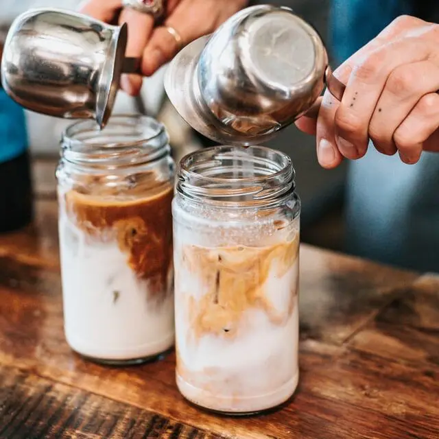 barista pouring milk into two iced lattes in mason jars on wood table, Are lattes hot or cold?
Iced lattes

