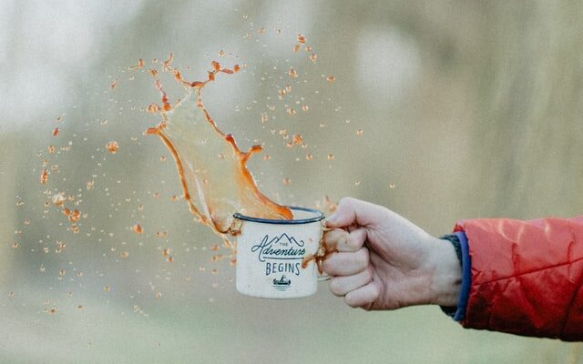 man's arm holding camping mug of coffee and splashing coffee up out of the mug, ground coffee vs instant coffee, instant coffee vs ground