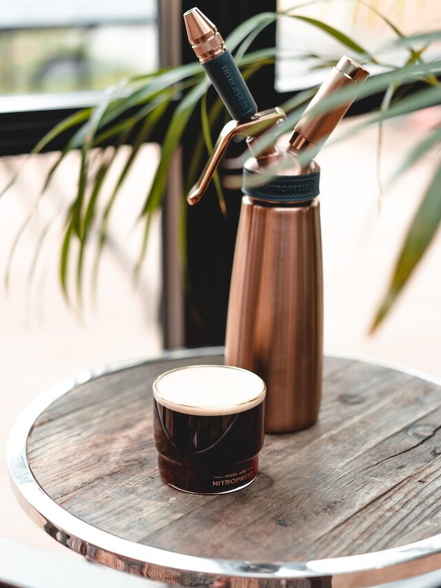 glass of nitro cold brew on wooden table beside nitropress cold brew bottle