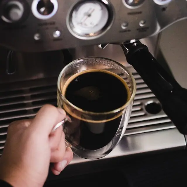 hand holding an americano coffee in a glass mug beside a Breville espresso machine